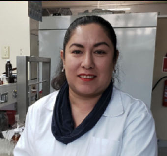 Mujer de complexión media, tez clara y cabello oscuro recogido, usando una bata blanca de laboratorio y un pañuelo oscuro alrededor del cuello, sonriendo ligeramente. Se encuentra en un laboratorio con equipos y estantes blancos de fondo.