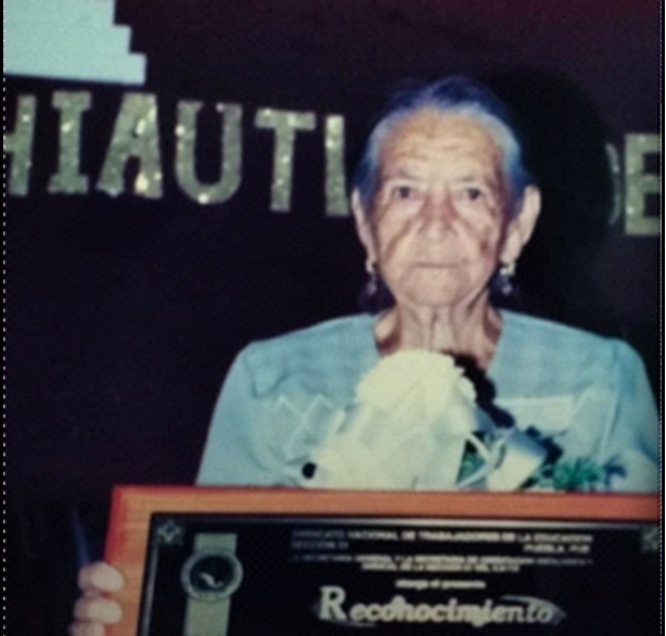 En la imagen se muestra aManuela Gertrudis Guerrero García, mejor conocida como Tulitas, quien fue pionera de la educación en Puebla. Ella se muestra en la imagen sosteniendo un reconocimiento, y unas flores blancas, con una ligera sonrisa. Ella  fue más que una maestra: fue una pionera de la educación en Puebla. A los 15 años comenzó a enseñar, y a lo largo de su vida, desafió barreras para que más niñas y niños tuvieran acceso al conocimiento.
