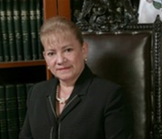 La imagen muestra un retrato de Zenorina González Ortega, quien esta en un espacio cerrado sentada y vestida con un traje negro, atrás se muestra un librero que parece ser un mueble que almacena libros del archivo historico, ella ha dedicado su vida a promover la igualdad de género y el empoderamiento de las mujeres. Como presidenta de la Comisión de Equidad de Género en la LVIII Legislatura del Congreso del Estado de Puebla (2011-2014), impulsó leyes fundamentales como la Ley de Acceso a las Mujeres a una Vida Libre de Violencia y la inclusión del feminicidio en el Código de Defensa Social.